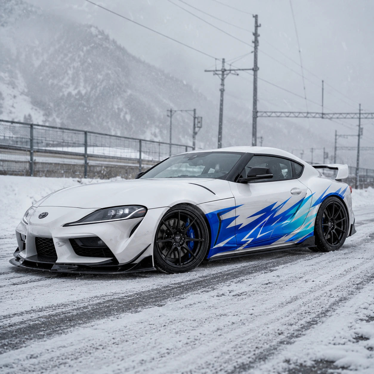 White Toyota Supra GR with blue lightning car side decal – front 3/4 view on snowy mountain road with overcast sky